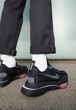 Personne portant des baskets Nike noires avec des chaussettes blanches et un pantalon noir retroussé, debout sur une surface en béton.