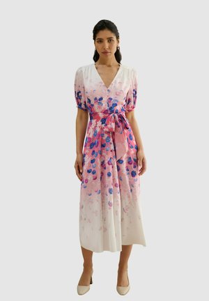 Mujer con vestido midi blanco y rosa con estampado floral, mangas cortas, escote en V, cinturón anudado en la cintura y zapatos de tacón beige.
