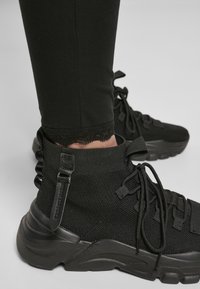 Zwarte high-top sneakers van textuur-mesh en rubber met veters en zijgespen. Draagt met strakke zwarte leggings.