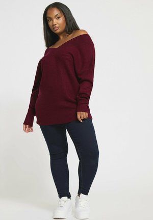 Burgundy yli suuri neulepaita, jossa on ribbiohennus ja olkapäiltä paljastava design, yhdistettynä tummiin slim-fit farkkuihin ja valkoisiin lenkkareihin.