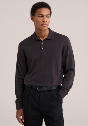 Jeune homme portant un polo à manches longues de couleur foncée, rentré dans un pantalon noir plissé avec une ceinture noire, debout, une main dans la poche.