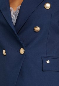 Laivastonsininen bleiseri, joka on valmistettu kuosisesta kankaasta, kultasävyisillä nappeilla ja yhdellä taskutiedoilla. Kukkalace-viimeistely näkyy kaula-aukossa.