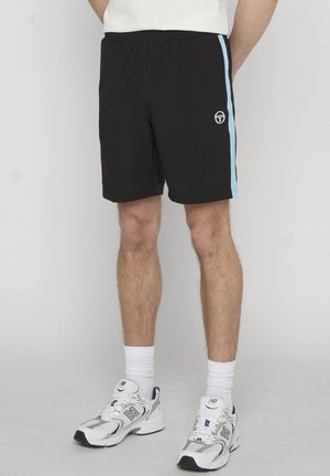 Mann trägt schwarze Sportshorts mit hellblauen Seitenstreifen und weiße Sneakers mit Socken, steht vor einem schlichten Hintergrund.