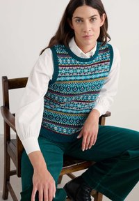 Gilet in maglia di colore teal con motivi geometrici, abbinato a una camicia bianca e pantaloni di velluto a coste verde scuro, seduto su una sedia di legno.
