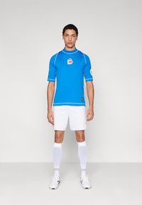 Modrý dres s krátkými rukávy s bílými akcenty a logem, doplněný bílými šortkami, bílými chrániči holení a kopačkami. Jednoduchý atletický design.