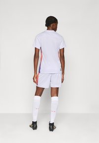 Weißes Fußballtrikot mit kurzen Ärmeln, verziert mit lila und korallenfarbenen Akzenten an den Seiten; kombiniert mit weißen Shorts und knielangen weißen Socken.