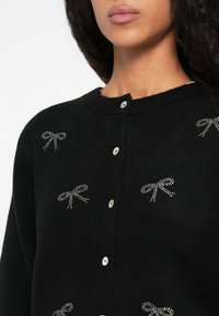Prim-plan al unui cardigan negru cu nasturi albi mici și embellismente cu fundițe din rhinestone argintii pe piept și umeri.