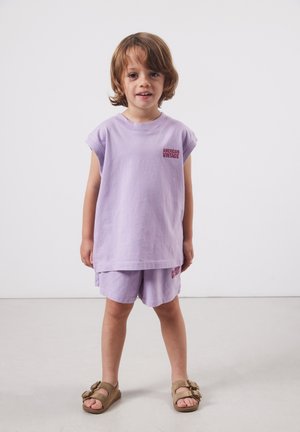 Mladé dieťa s vlasmi po plecia, oblečené v svetlofialovej tielkovej blúzke a šortkách s béžovými sandálmi, stojace na jednoduchom pozadí.