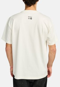 Cream-colored short-sleeve T-shirt with a relaxed fit and a small black logo on the upper back. Soft fabric with a smooth texture.