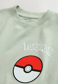 Mintgrøn sweater med et rundt Pokémon-logo og hvid tekst. Halsudskæringen er ribbet, og stoffet har en glat, blød tekstur.