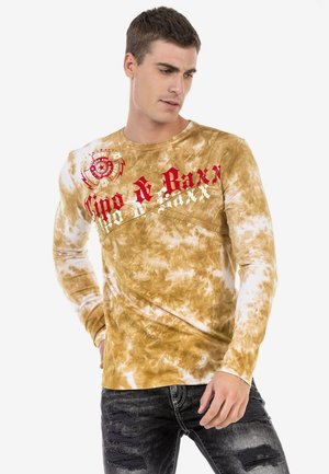Langärmliges T-Shirt mit Batikmuster in Braun und Beige, das rote und weiße Schriftzüge aufweist. Der Stoff hat eine weiche, strukturierte Optik mit einem runden Ausschnitt.