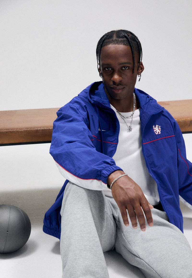 Blå nylonjakke med røde detaljer, hvit kant og hette; grå joggebukser; sølvsmykker; sitter på benk med en grå ball i nærheten.