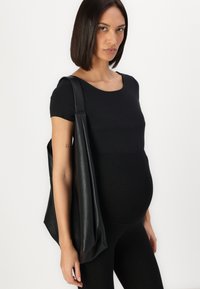 Črna usnjena torba s teksturirano površino, nošena čez prilegajočo se črno majico in legice. Model ima zaobljen trebuh.