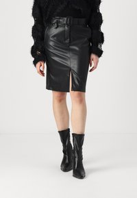 Μαύρη ψεύτικη δερμάτινη μίντι φούστα με μπροστινή σχισμή, ζωνάκι στη μέση και λεία υφή, συνδυασμένη με μαύρες δερμάτινες μπότες μέχρι το γόνατο.