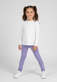 Biała koszula z długim rękawem w połączeniu z lawendowymi legginsami; modelka nosi białe trampki; gładka tekstura i dopasowany fason na całej powierzchni.