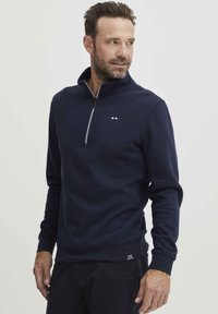 Polo majica u tamnoplavoj boji s četvrtastim patentnim zatvaračem, izrađena od mekane tkanine, s ovratnikom na patentni zatvarač i malim logotipom blizu grudi.