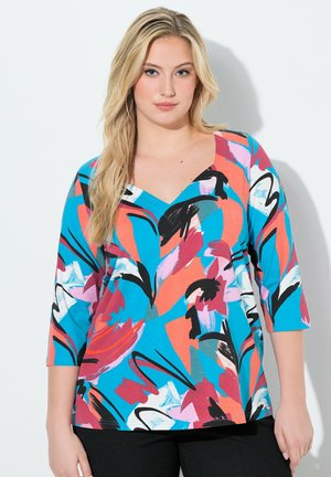 Blonde Frau trägt ein V-Ausschnitt-Oberteil mit dreiviertellangen Ärmeln und markanten abstrakten Pinselstrichmustern in Blau, Koralle, Schwarz, Pink und Weiß.
