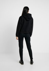 Mujer vestida con una sudadera negra, jeans ajustados negros y zapatos negros, de pie de espaldas contra un fondo blanco liso.