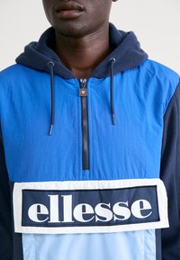 Blå och marinblå huva jacka med halvdol zip-fram, med en stor vit och marinblå logotyp patch. Strukturerad tygkontrast vid axlarna.