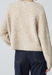 Beige fuzzy sweater with a loose fit, textured fabric, and ribbed cuffs. Paired with dark blue jeans featuring subtle stitching detail.