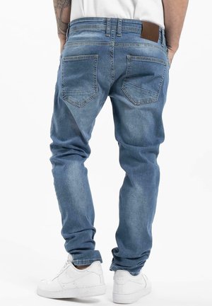 Person iført lyseblå denimjeans og hvide sneakers vist bagfra, stående med hænderne i lommerne mod en hvid baggrund.