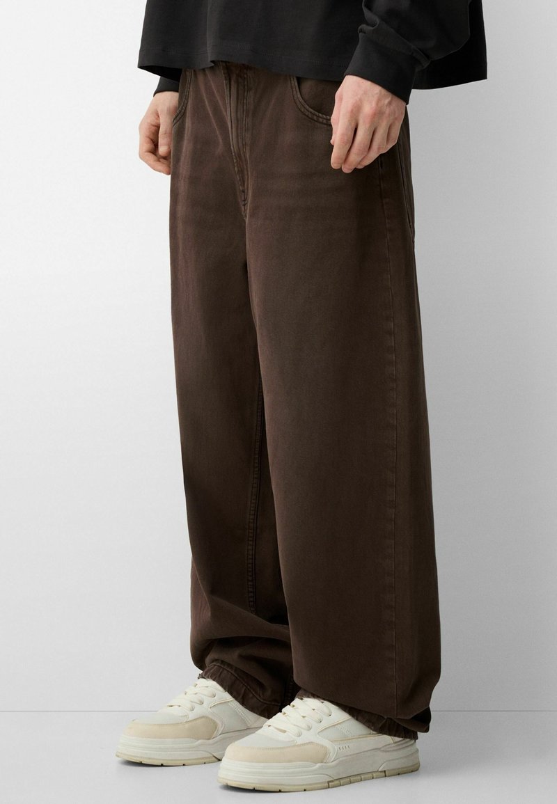 Pantalones anchos marrones de algodón, con una textura suave, lazos para cinturón y bolsillos delanteros, combinados con zapatillas de deporte crema.