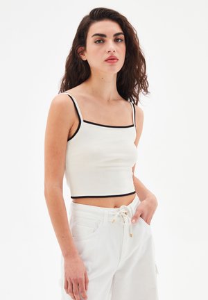 Bílý kratší top s obdélníkovým výstřihem a tenkým černým lemem. Modelka ho kombinuje s bílými kalhotami na šňůrku. Hladká textura látky.