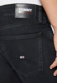Acercamiento al bolsillo trasero de unos jeans de mezclilla negra con un pequeño logo de Tommy Jeans y una etiqueta de cuero de la marca encima de la cinturilla.