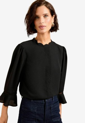 Žena s krátkymi hnedými vlasmi, ktorá má zlaté kruhové náušnice, čiernu blúzku s vlnitým golierom a riasenými rukávmi a tmavomodré nohavice s vysokým pásom.