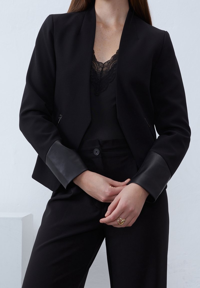 Blazer nero con una texture liscia, dotato di dettagli in pelle sui polsini. Indossato sopra un top di pizzo nero, abbinato a pantaloni neri.