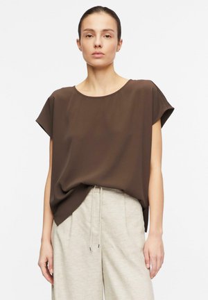 Donna che indossa una blusa marrone morbida a maniche corte infilata in pantaloni beige chiaro con coulisse, in piedi davanti a uno sfondo bianco semplice.