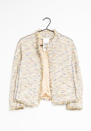 Giacca corta in tweed multicolore con bordi sfrangiati e bottoni frontali, appesa a una gruccia nera su sfondo bianco.