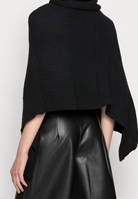 Vue arrière d'une personne portant une cape en maille noire avec de larges ouvertures aux bras et un pantalon en cuir noir taille haute, sur fond blanc.