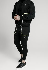 Chándal deportivo negro con acentos neón, que incluye una chaqueta con cremallera y pantalones ajustados. Logo metálico en el pecho y bolsillos laterales. Zapatillas negras.