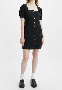 Musta farkku mekko, jossa on puffihihat, neliskulmainen kaula-aukko ja etupainonappi. Mukana on istuva vyötärö ja polven ylle ulottuva pituus.