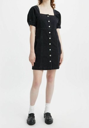 Musta farkku mekko, jossa on puffihihat, neliskulmainen kaula-aukko ja etupainonappi. Mukana on istuva vyötärö ja polven ylle ulottuva pituus.