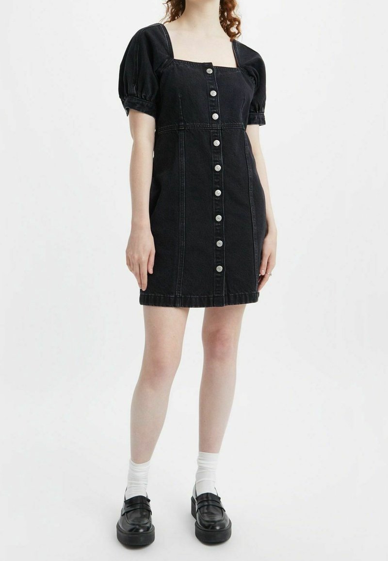 Musta farkku mekko, jossa on puffihihat, neliskulmainen kaula-aukko ja etupainonappi. Mukana on istuva vyötärö ja polven ylle ulottuva pituus.