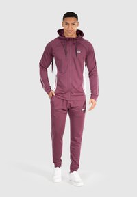 Bordeaux tracksuit med hvide sidelommer, der inkluderer en zip-up hoodie og matchende bukser, lavet af glat, strækbart materiale. Hvide sneakers.