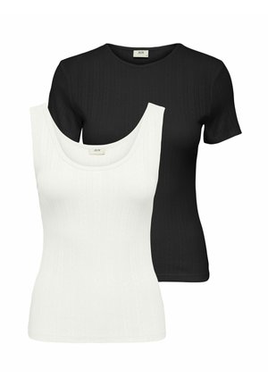 Femme debout portant un top noir ajusté à manches courtes, un pantalon noir large taille haute et des chaussures blanches pointues sur un fond uni.