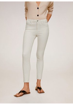 Woman wearing beige and white vertical striped skinny pants and beige cardigan, standing with hands in pockets and black sandals.