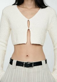 Cardigan scurt alb din tricot, cu decolteu în V adânc, decupaj central proeminent și mâneci lungi, asortat cu pantaloni gri cu dungi și talie înaltă, purtând o curea neagră.