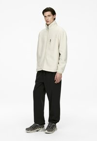 Light beige fleece jacket with a high collar and zip closure, featuring a chest pocket. Paired with loose-fitting black trousers and gray sneakers.