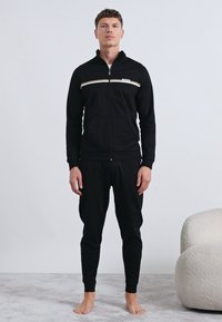 Μαύρη φόρμα με ζακέτα με φερμουάρ, που έχει οριζόντια λευκή και χρυσή ρίγα, και ταιριαστό παντελόνι. Κατασκευασμένη από μαλακό, ελαστικό ύφασμα.