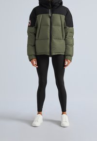 Person wearing an olive green and black puffer jacket, black leggings, and white sneakers standing against a plain light background.