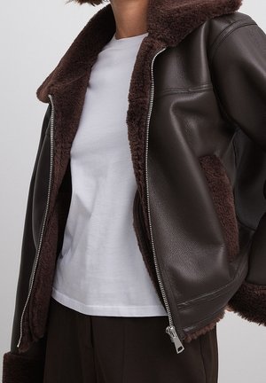 Personne portant une veste en cuir marron foncé avec une doublure en shearling, partiellement zippée sur un t-shirt blanc et un pantalon foncé.