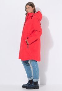 Czerwona wodoodporna parka z kapturem, wyposażona w przedni zamek błyskawiczny, kieszenie na zamek i czarne akcenty. Nosi się ją na spodniach z podwiniętymi nogawkami i ciemnych butach.