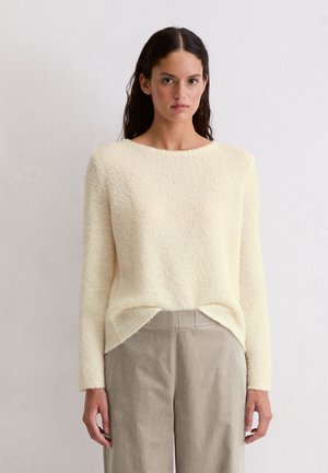 Vrouw met lang donker haar draagt een crèmekleurige gebreide trui en losse beige broek, staande tegen een effen lichte muur.