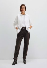 Camicia bianca a bottoni sopra una t-shirt bianca, abbinata a pantaloni di velluto a coste marrone scuro risvoltati e stivaletti neri. Tessuti lisci e vestibilità sartoriale.