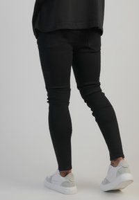 Schwarze Slim-Jeans aus Denim, mit einer glatten Textur und ausgefranstem Design. Getragen mit weißen Sneakern mit grauen Akzenten.