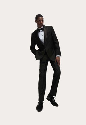 Czarny smoking z satynowymi klapami, biała koszula z czarnymi guzikami oraz czarne buty. Garnitur jest dopasowany, nadając ostry, wyraźny kształt sylwetce.